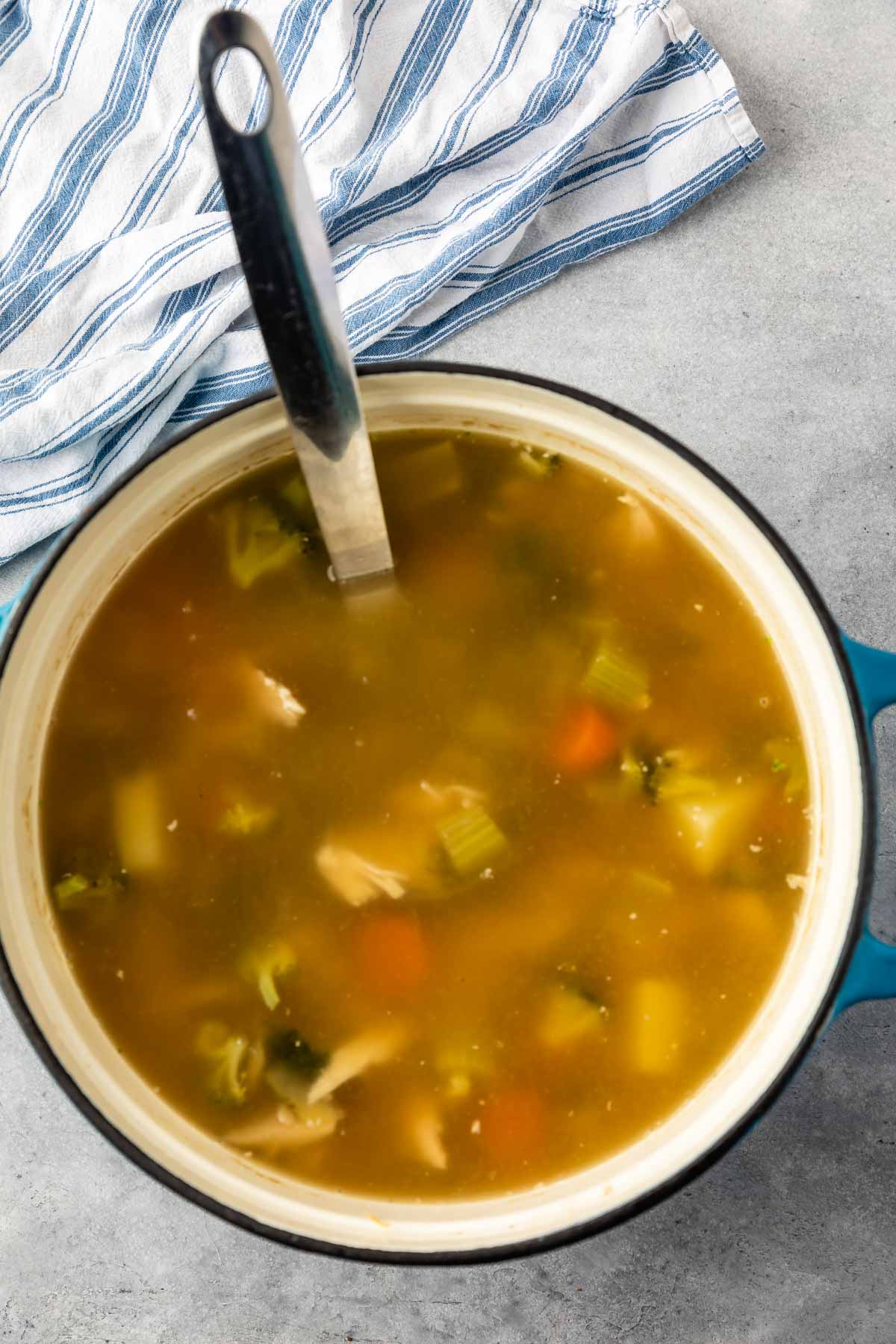 A large blue pot filled with vegetable and chicken soup sits on a gray surface, with a ladle inside. A blue and white striped cloth is partially visible in the background.