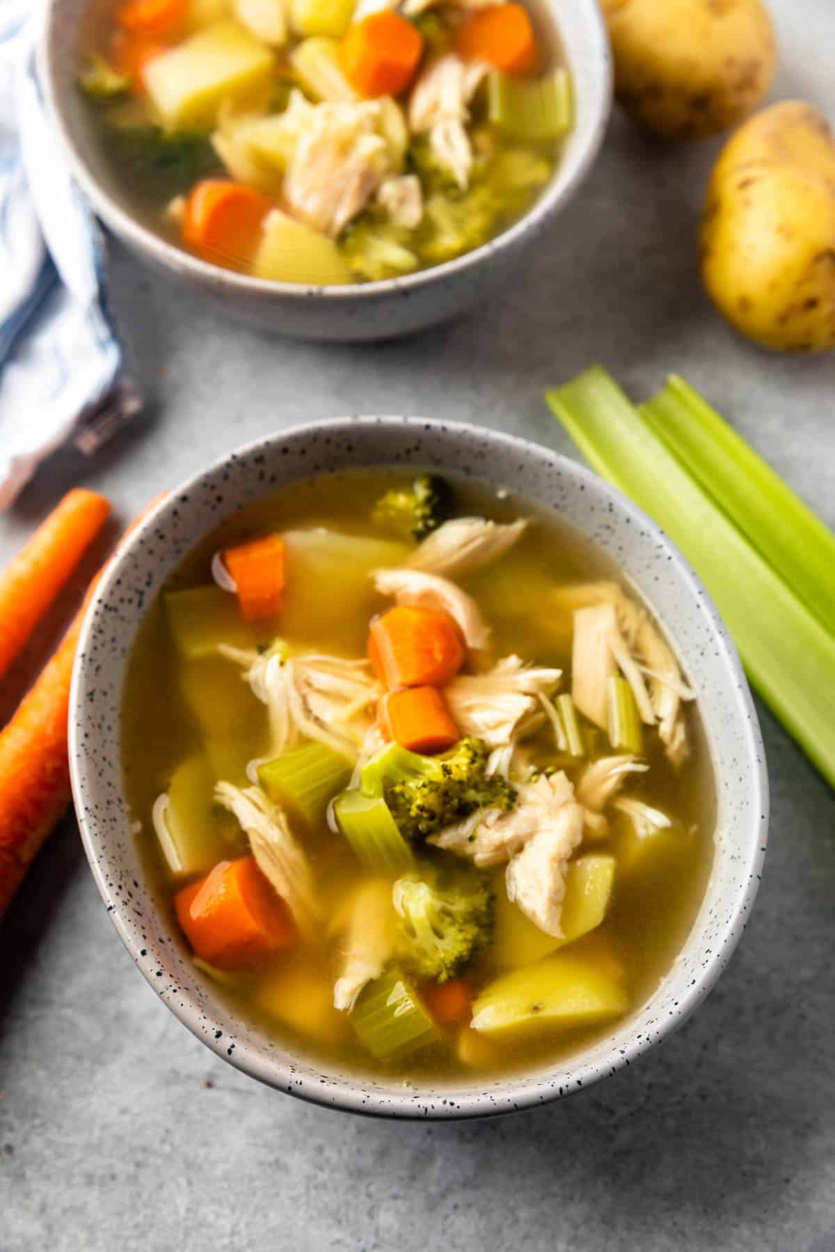 A bowl of chicken vegetable soup filled with carrots, celery, broccoli, potatoes, and shredded chicken sits on a gray surface alongside fresh celery stalks, carrots, and potatoes. Another bowl of soup is in the background.