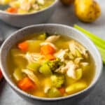 A bowl of chicken vegetable soup with carrots, celery, broccoli, and shredded chicken in broth, with potatoes and a second bowl of soup in the background.
