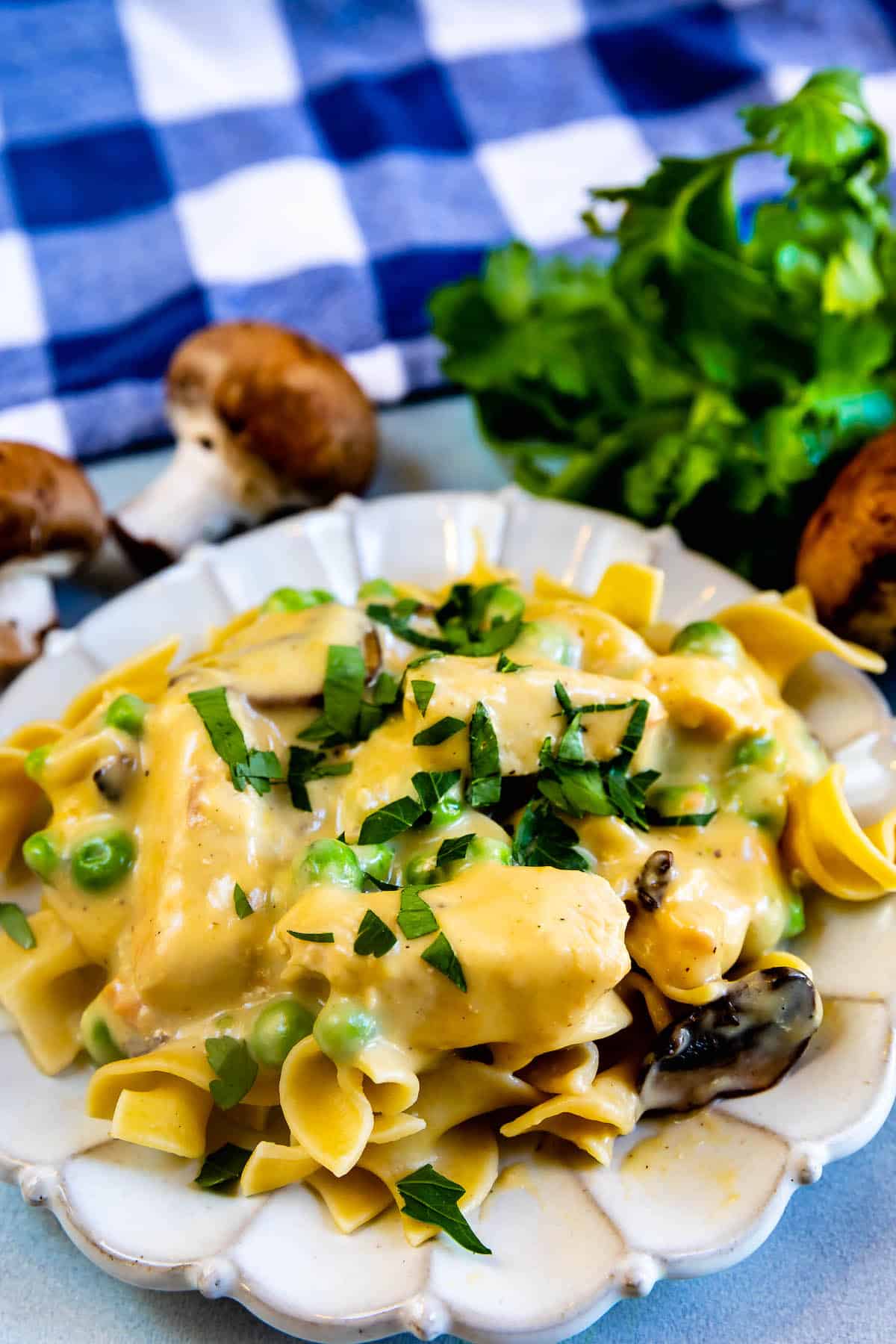 A plate of creamy pasta with peas, mushrooms, and fresh parsley sits on a white dish. In the background, a bunch of parsley, whole mushrooms, and a blue-and-white checkered cloth are visible.