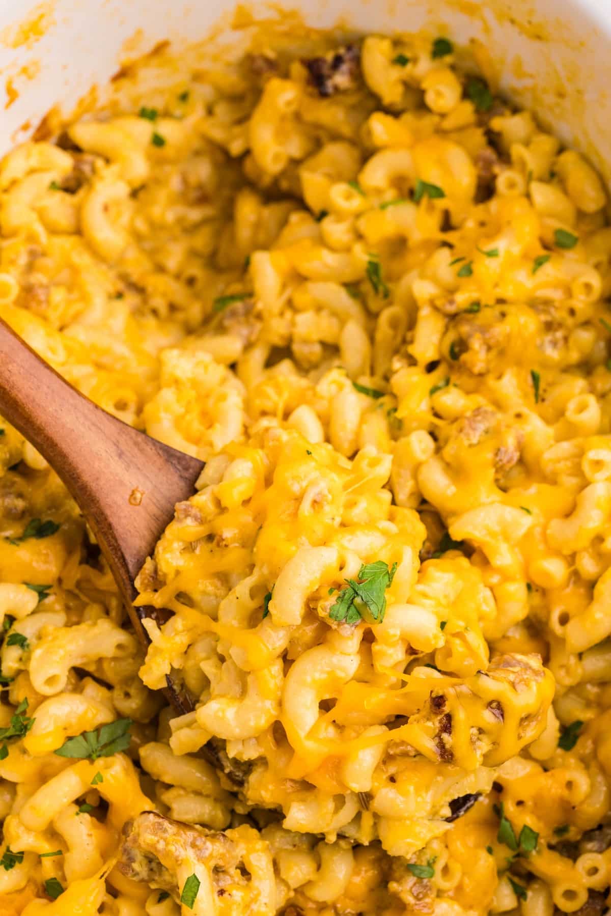 Creamy macaroni and cheese with melted cheddar, ground meat, and garnished with chopped parsley, being scooped with a wooden spoon.