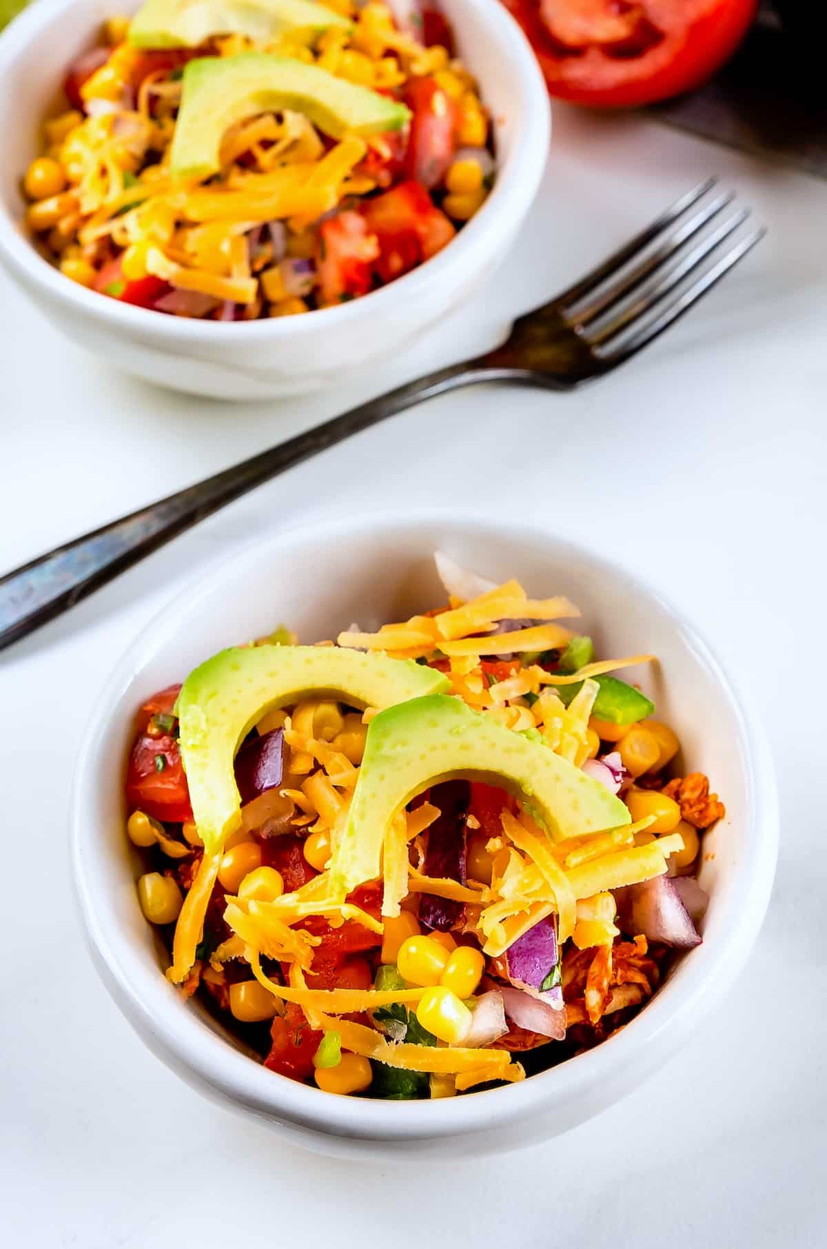 A white bowl filled with a colorful salad featuring corn, diced tomatoes, red onion, shredded cheddar cheese, and avocado slices, with a fork and another bowl of salad in the background.