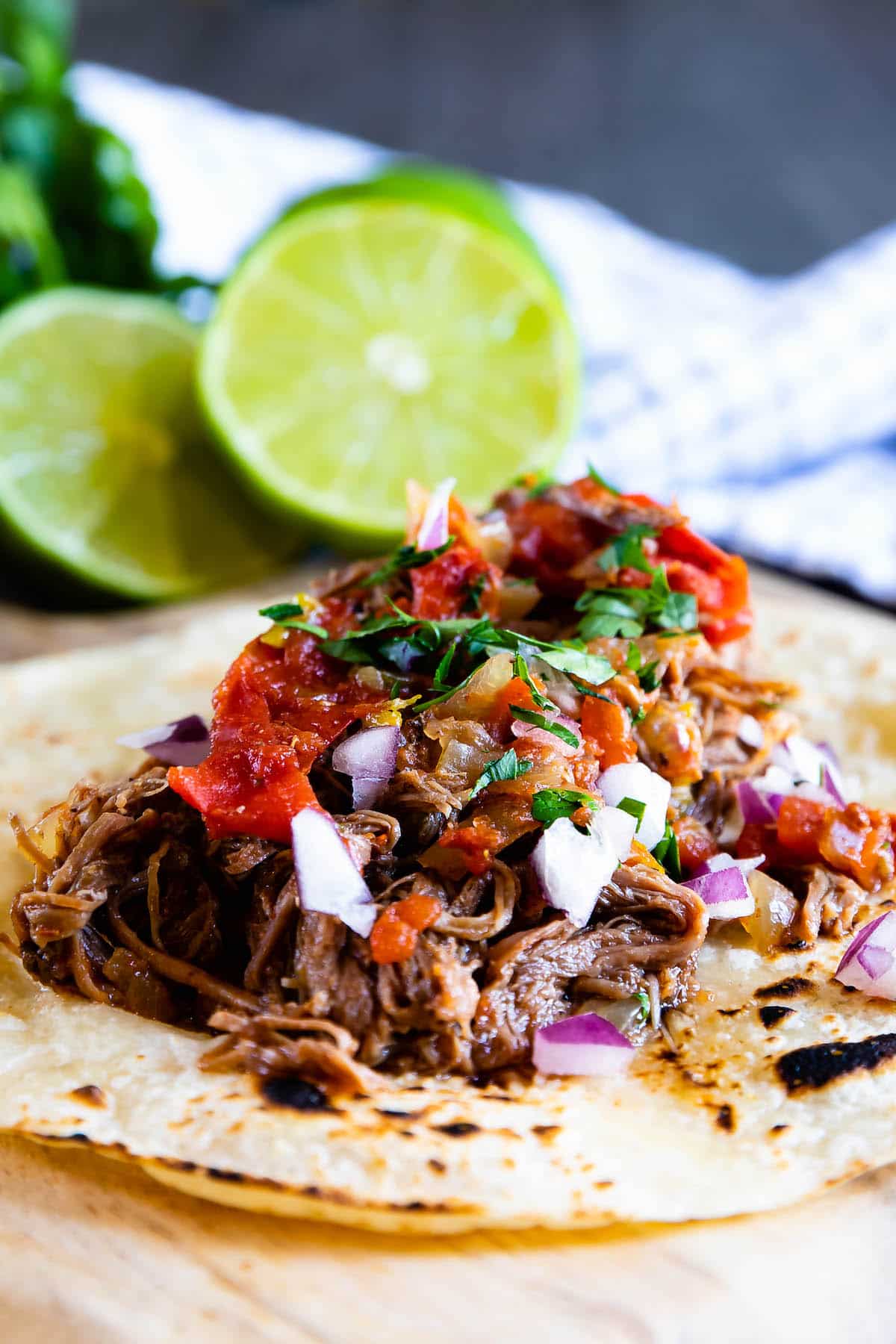A close-up of a taco with shredded beef, diced red onions, fresh salsa, and chopped cilantro on a toasted tortilla. Sliced limes and a cloth napkin are in the blurred background.