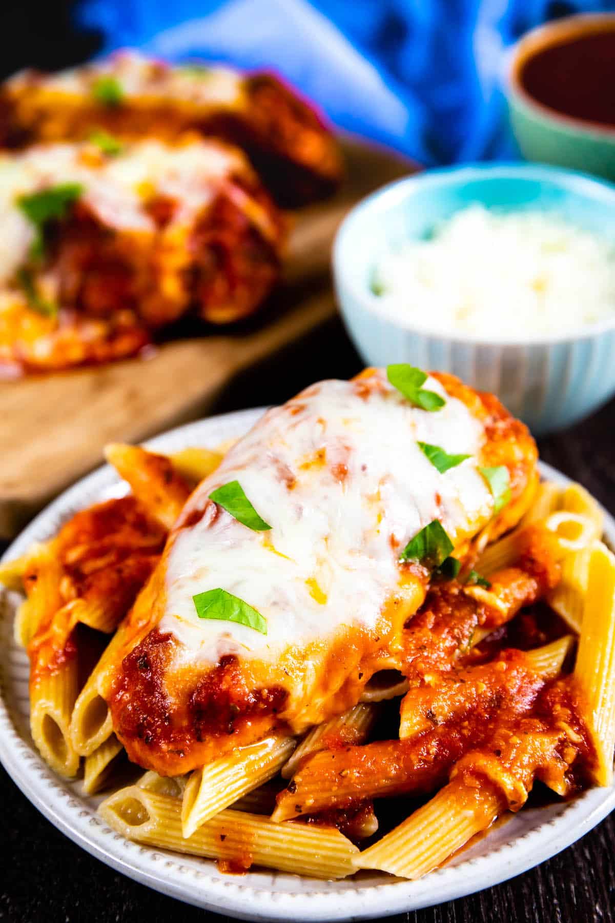 A plate of penne pasta topped with chicken parmesan, melted cheese, tomato sauce, and fresh basil. In the background, more chicken parmesan, grated cheese, and a bowl of sauce are visible.