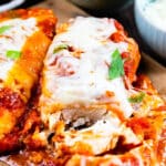 Close-up of cheesy baked chicken parmesan topped with tomato sauce and herbs, served on a wooden board, with a side of grated cheese and extra sauce in the background.