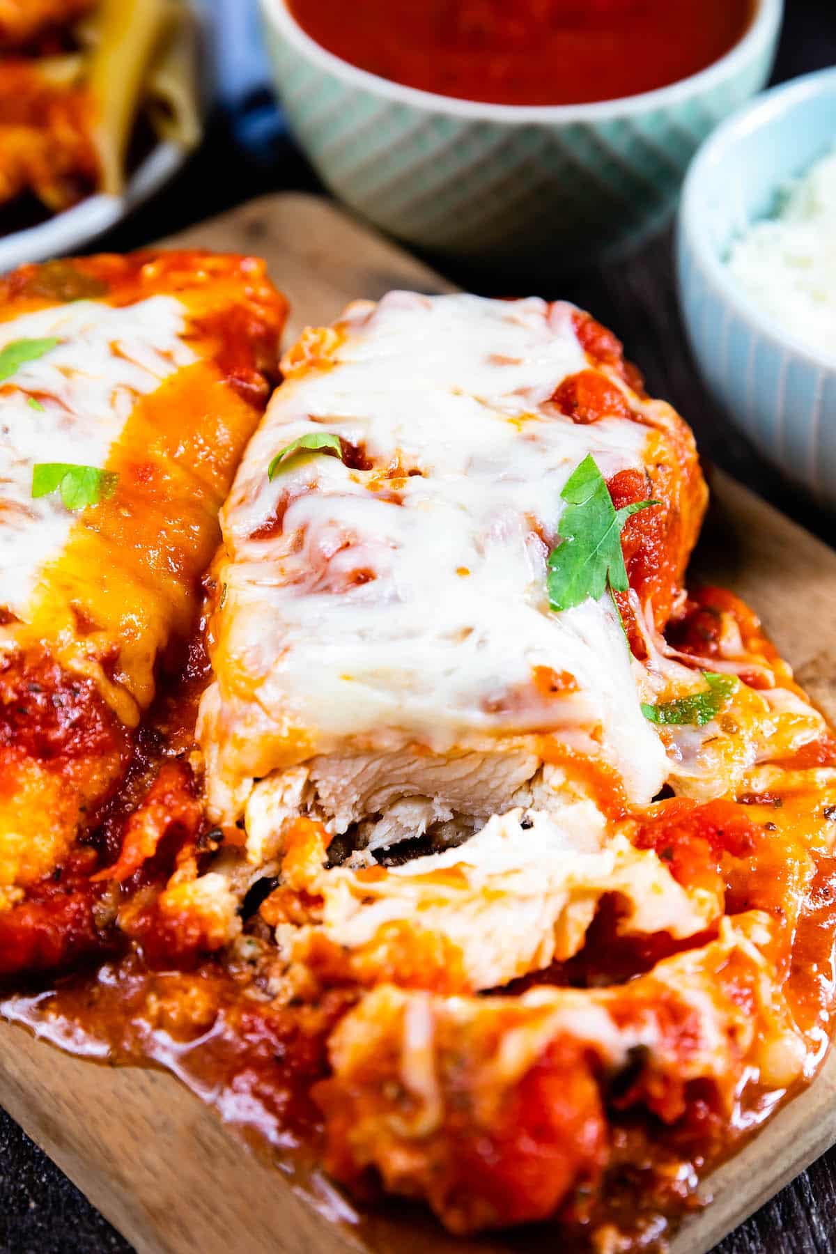 Close-up of cheesy baked chicken parmesan topped with tomato sauce and herbs, served on a wooden board, with a side of grated cheese and extra sauce in the background.