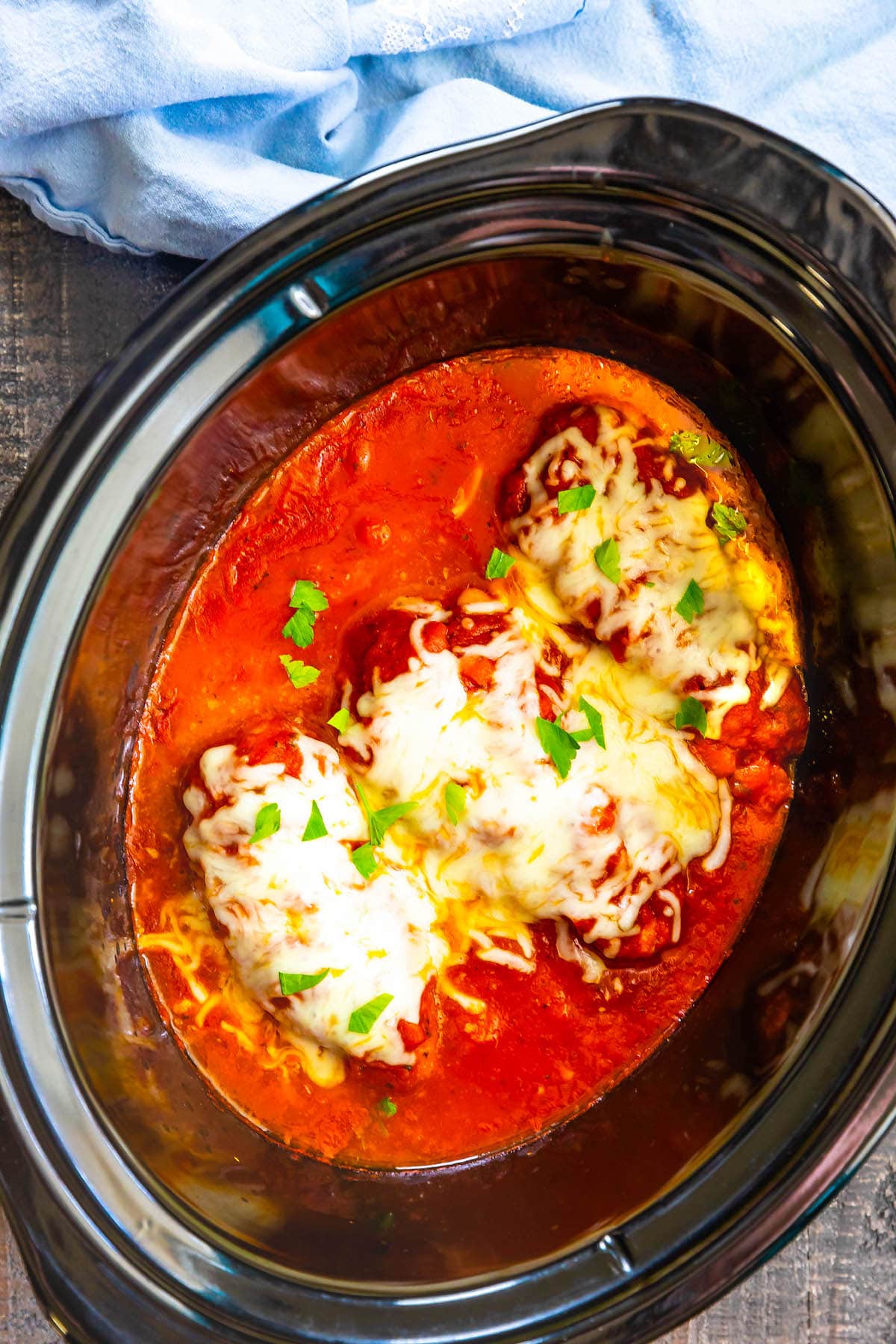 Three chicken breasts topped with melted cheese and parsley sit in a rich red tomato sauce inside a black slow cooker, with a blue cloth in the background.