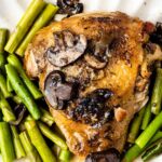 A cooked chicken thigh topped with mushrooms, served on a white plate with sautéed asparagus spears and a light sauce.
