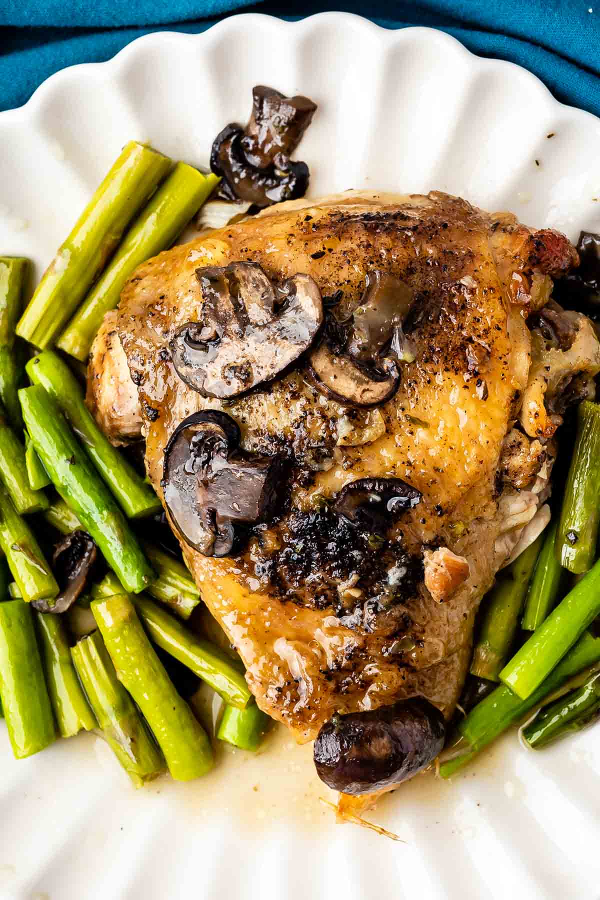 A cooked chicken thigh topped with mushrooms, served on a white plate with saut&eacute;ed asparagus spears and a light sauce.