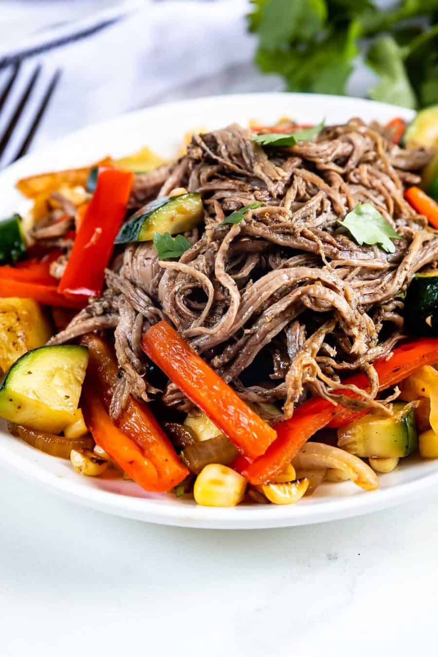 A plate of shredded beef served on top of sautéed vegetables including zucchini, red bell peppers, and corn, garnished with fresh herbs.