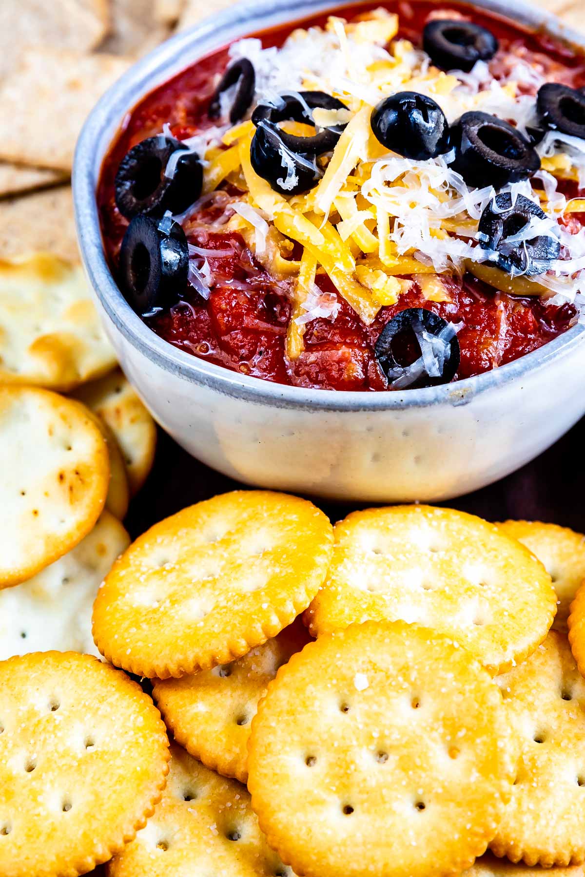 A bowl of chunky dip topped with shredded cheese and sliced black olives, surrounded by round, golden crackers and some irregularly shaped chips.