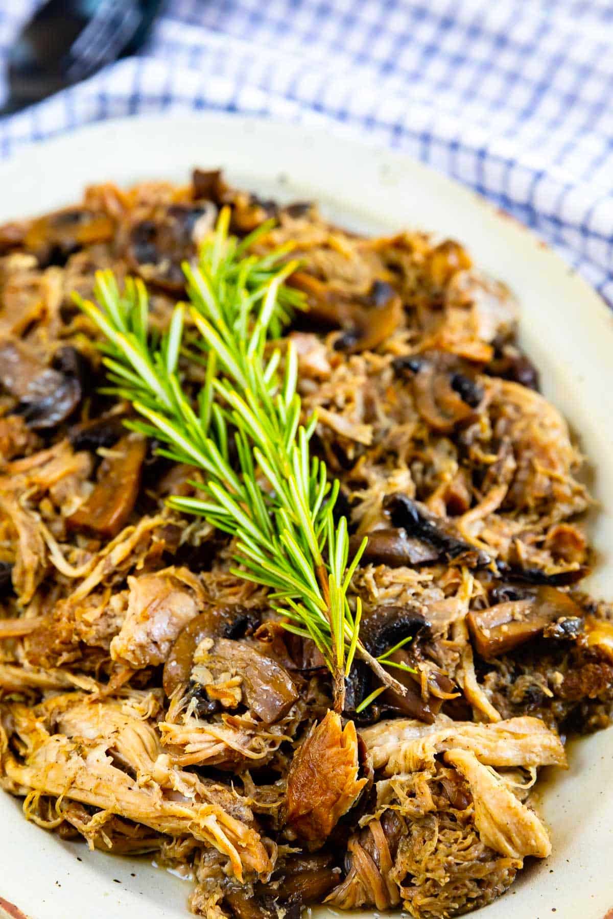A plate of shredded cooked meat mixed with mushrooms, garnished with a sprig of fresh rosemary, served on a white dish with a blue and white checkered cloth in the background.