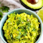 A bowl of fresh guacamole topped with chopped cilantro, with a halved avocado and lime wedge in the background.