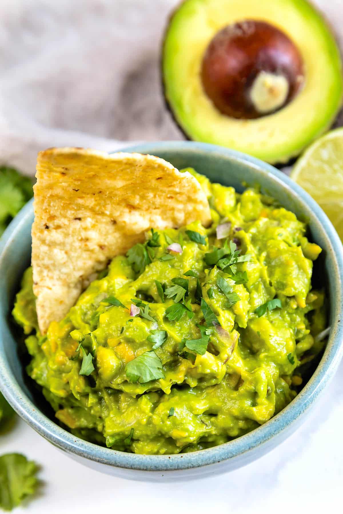 A bowl of guacamole topped with chopped cilantro, with a tortilla chip dipped in it. In the background, there is a halved avocado and a lime.