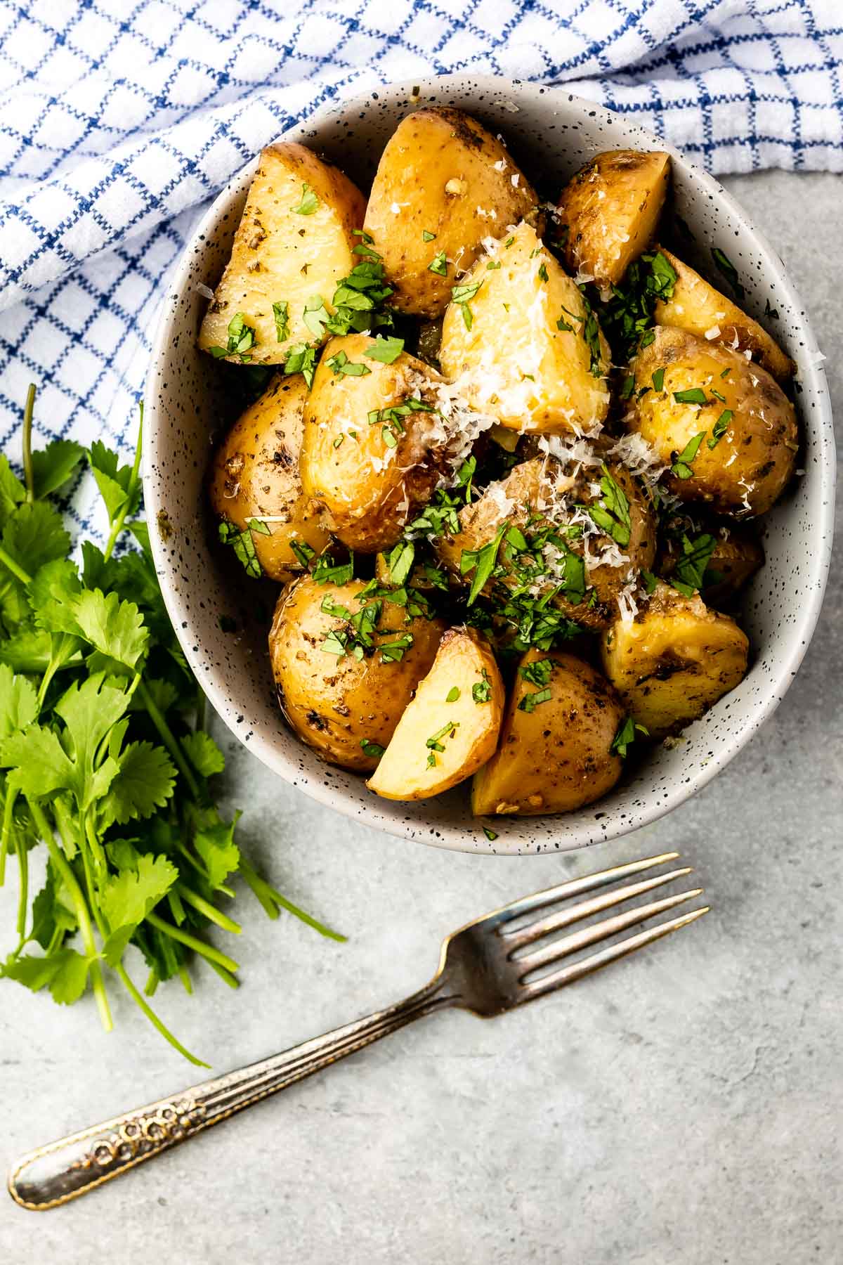 A bowl of seasoned roasted potato wedges topped with grated cheese and chopped herbs sits on a gray surface beside a fork, a bunch of fresh cilantro, and a blue-checked cloth napkin.