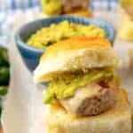 A slider sandwich with melted cheese and guacamole is served on a soft bun, placed on parchment paper. In the background, more sliders and a blue bowl of guacamole are visible.