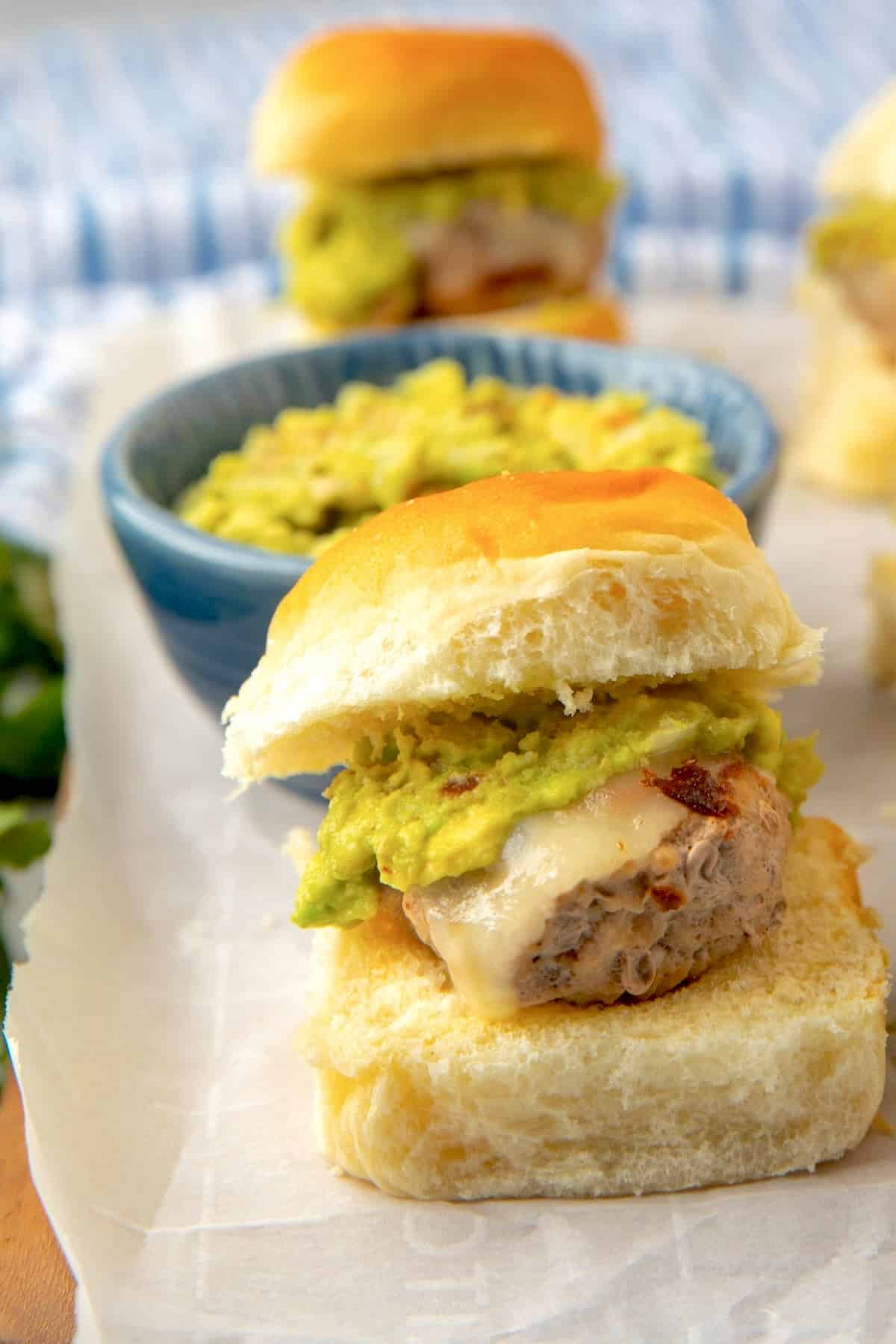 A slider sandwich with melted cheese and guacamole is served on a soft bun, placed on parchment paper. In the background, more sliders and a blue bowl of guacamole are visible.