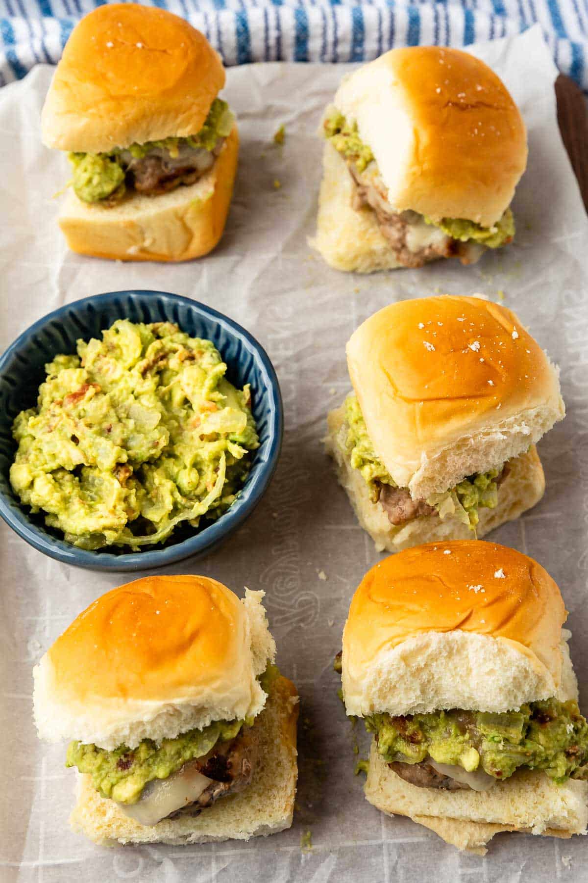 Five sliders filled with hamburger patties, melted cheese, and guacamole are arranged on parchment paper. A small blue bowl of extra guacamole sits among them. The buns look soft and golden brown.