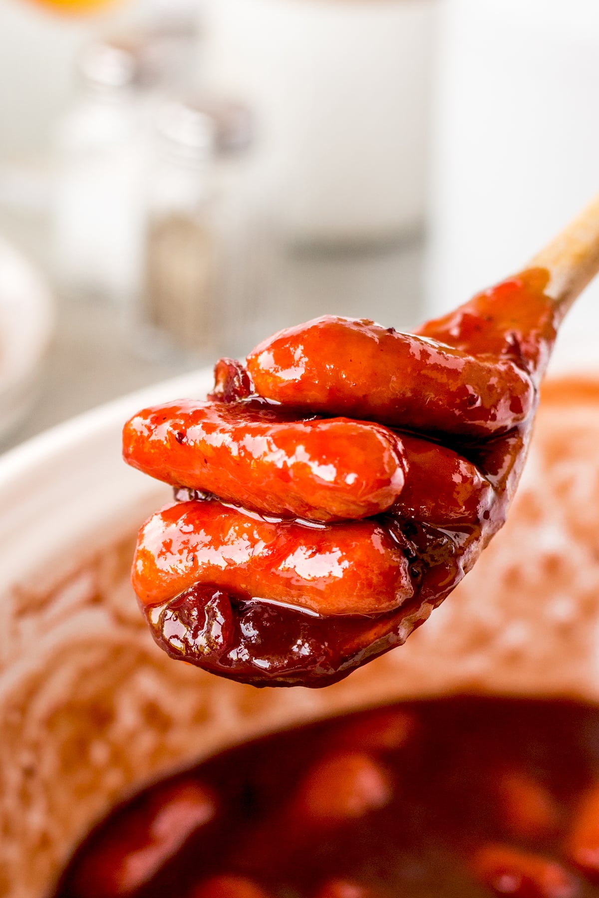 A wooden spoon holds several cocktail sausages coated in a thick, glossy reddish-brown sauce, with more sausages and sauce visible in a white bowl below.