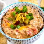 A bowl of creamy cheese dip with ground meat, diced tomatoes, and green onions, topped with shredded cheese, sliced jalapeños, and more green onions, surrounded by tortilla chips.