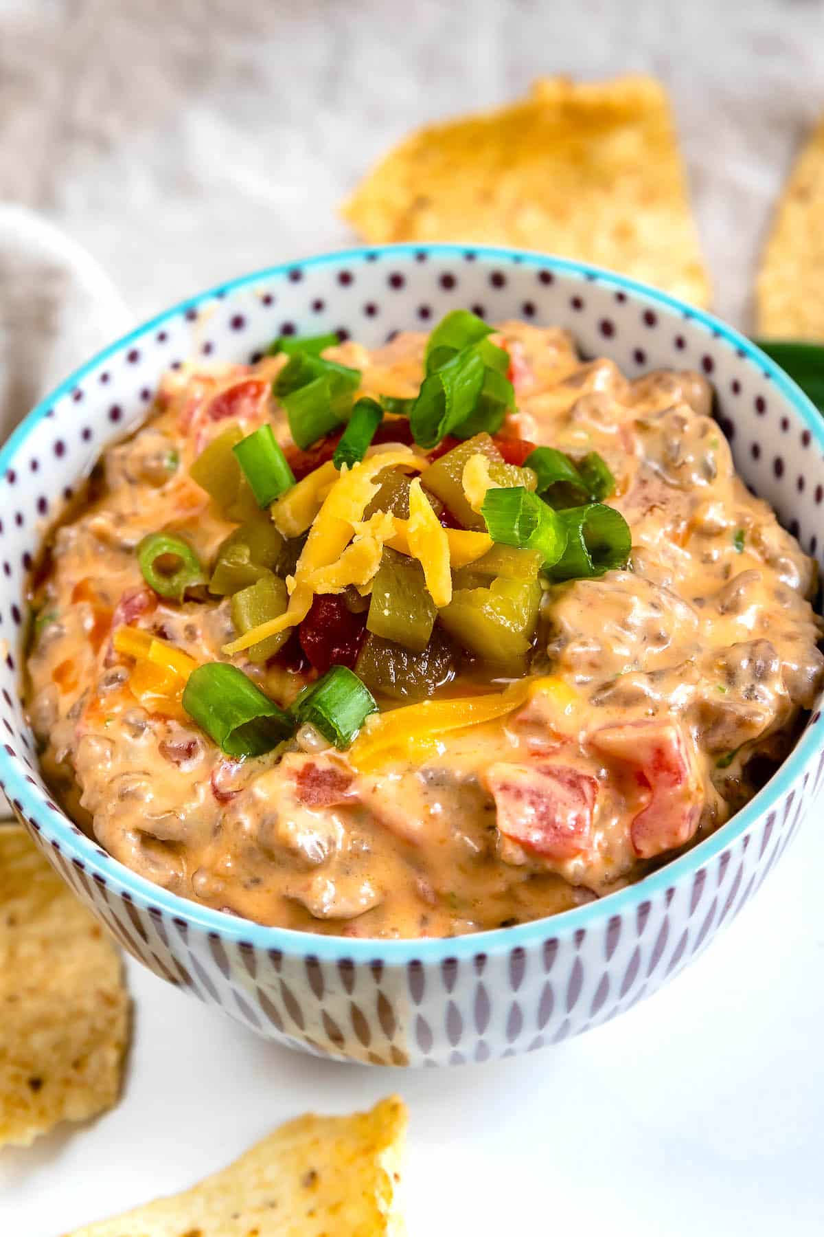 A bowl of creamy cheese dip with ground meat, diced tomatoes, and green onions, topped with shredded cheese, sliced jalapeños, and more green onions, surrounded by tortilla chips.