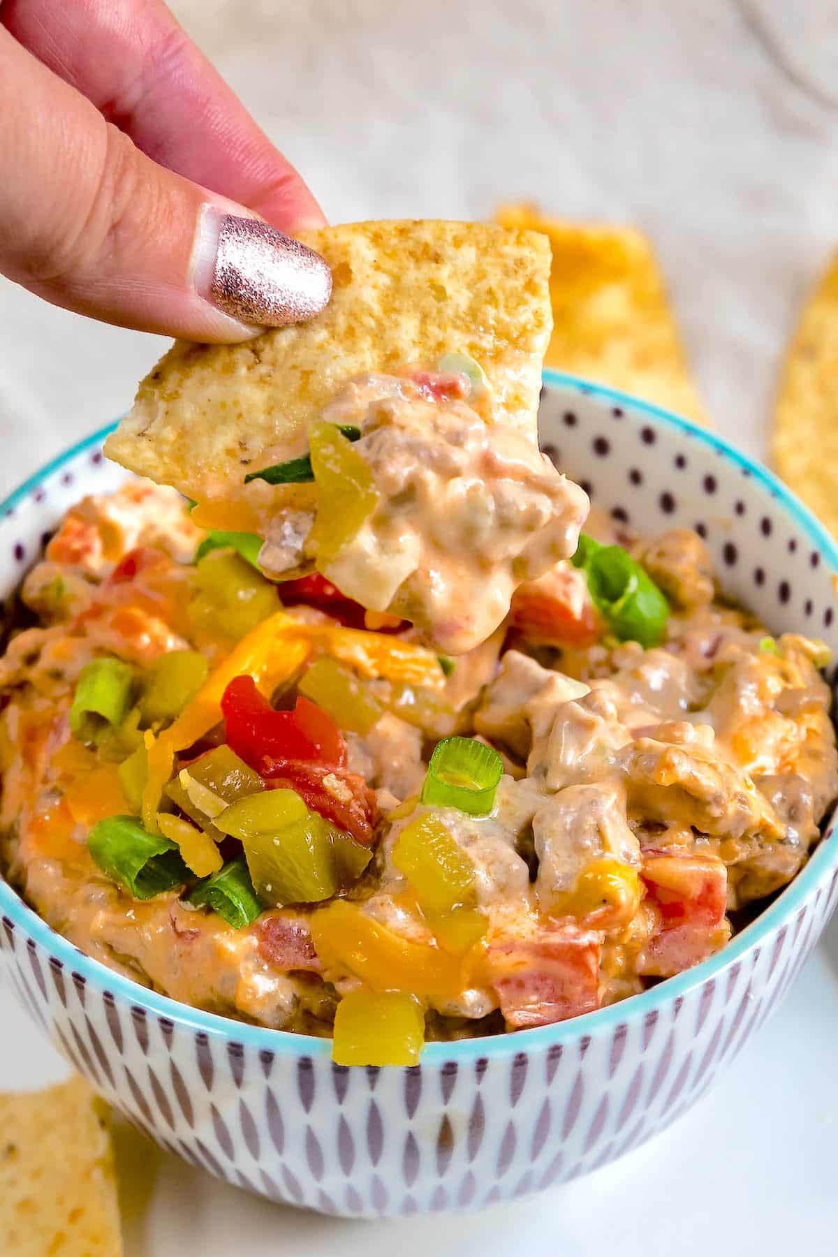 A hand holds a tortilla chip dipped in a creamy cheese and vegetable dip, garnished with chopped green onions and diced peppers, in a patterned bowl on a white plate.