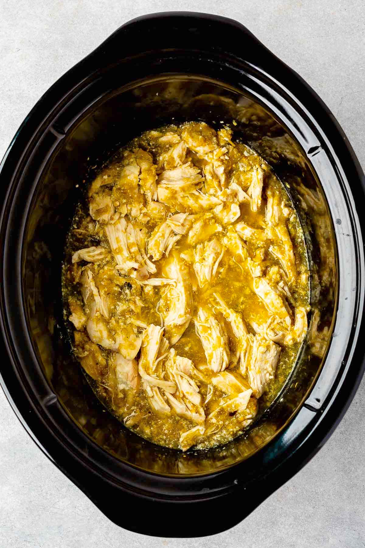 Shredded chicken in a yellowish sauce sits inside a black slow cooker, ready to be served.