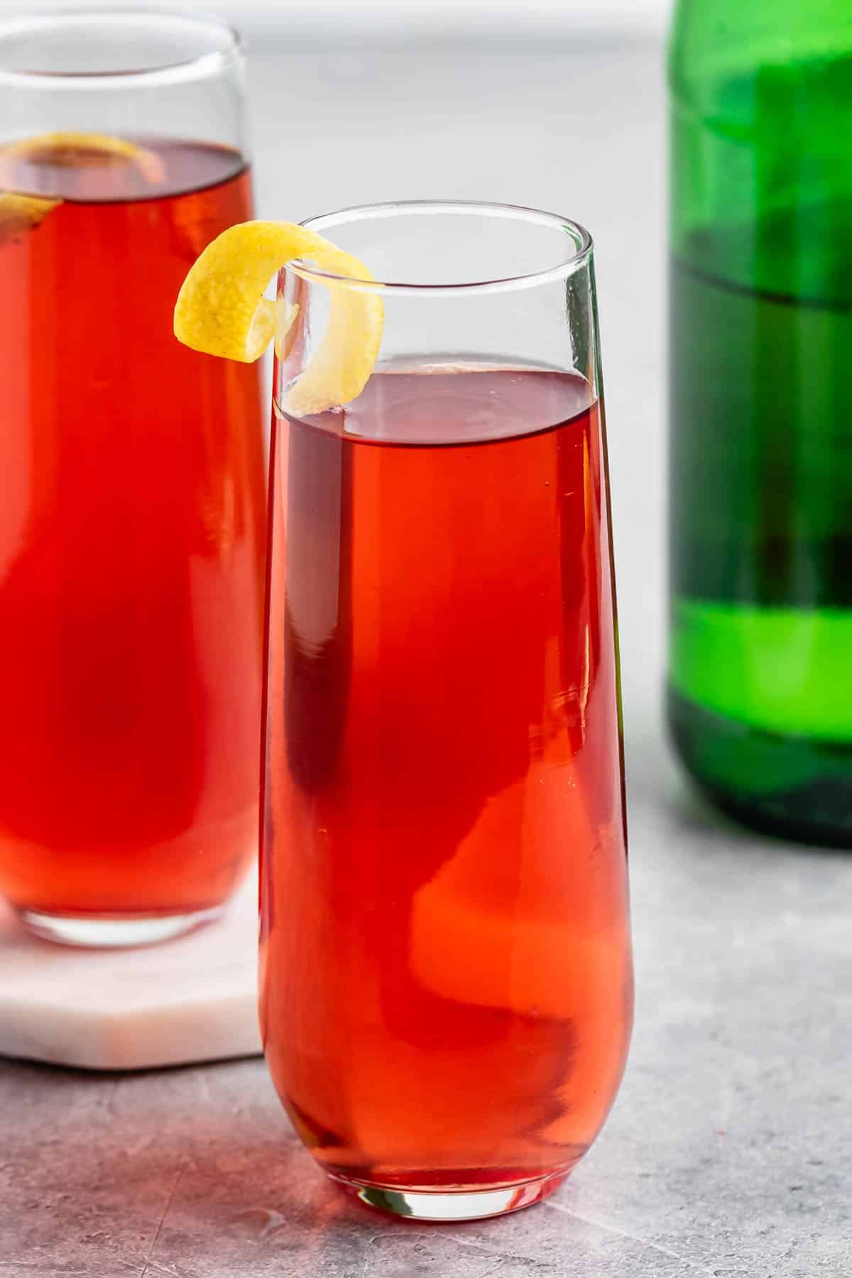 A tall glass filled with a red cocktail, garnished with a twist of lemon on the rim. Another similar glass and a green bottle are in the background on a light gray surface.