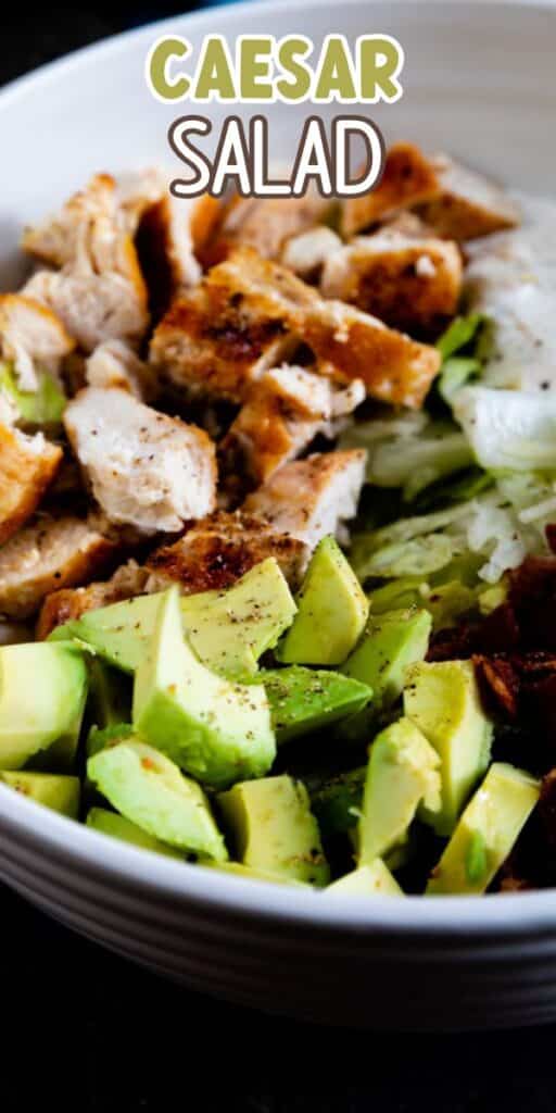 A bowl of Caesar salad topped with grilled chicken, avocado chunks, and lettuce. The text Caesar Salad is written at the top in bold, colorful letters.