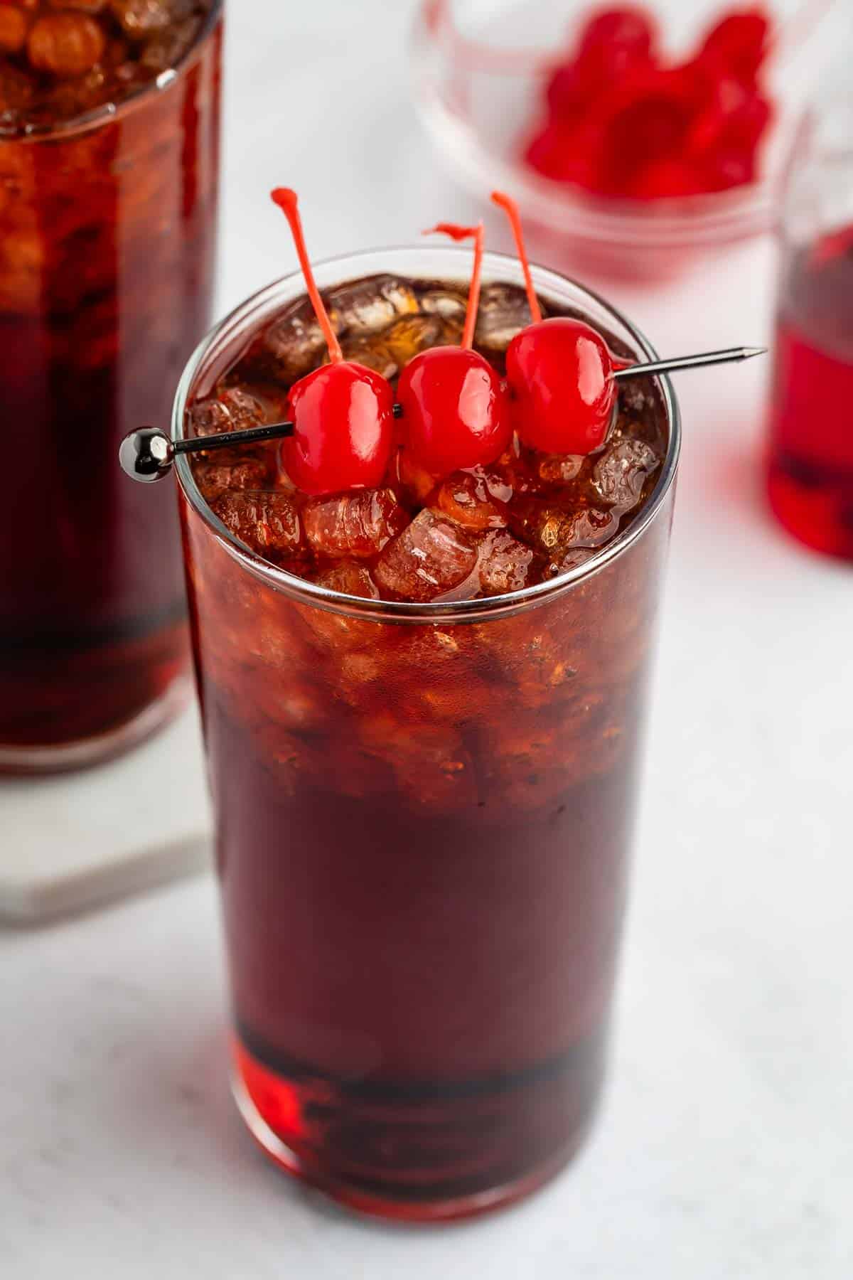 A tall glass filled with a dark, iced drink is garnished with three red maraschino cherries on a skewer. In the background, there is a bowl of cherries and another glass with a similar drink.
