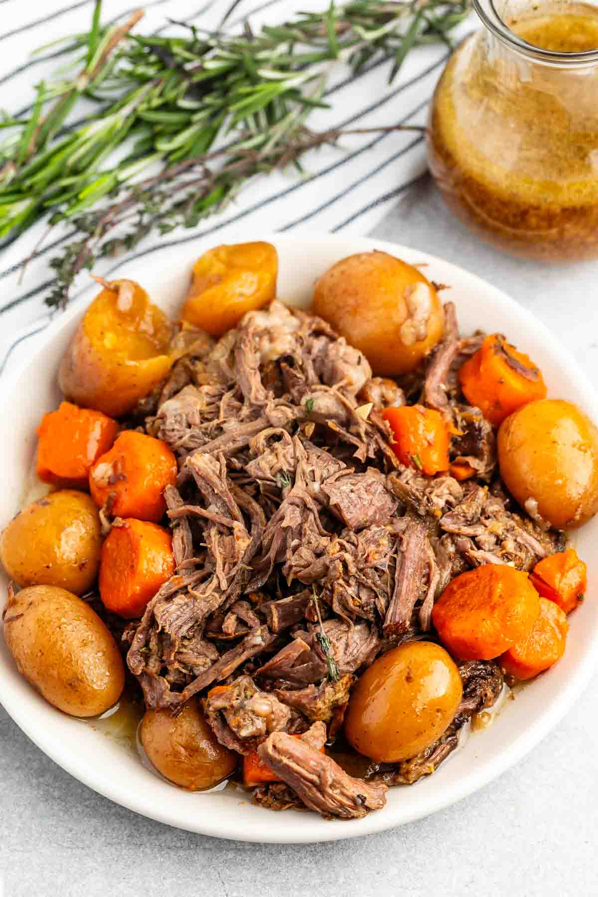 A white bowl filled with shredded pot roast, whole baby potatoes, and large carrot pieces, garnished with herbs. A jar of brown gravy and fresh rosemary and thyme are in the background.