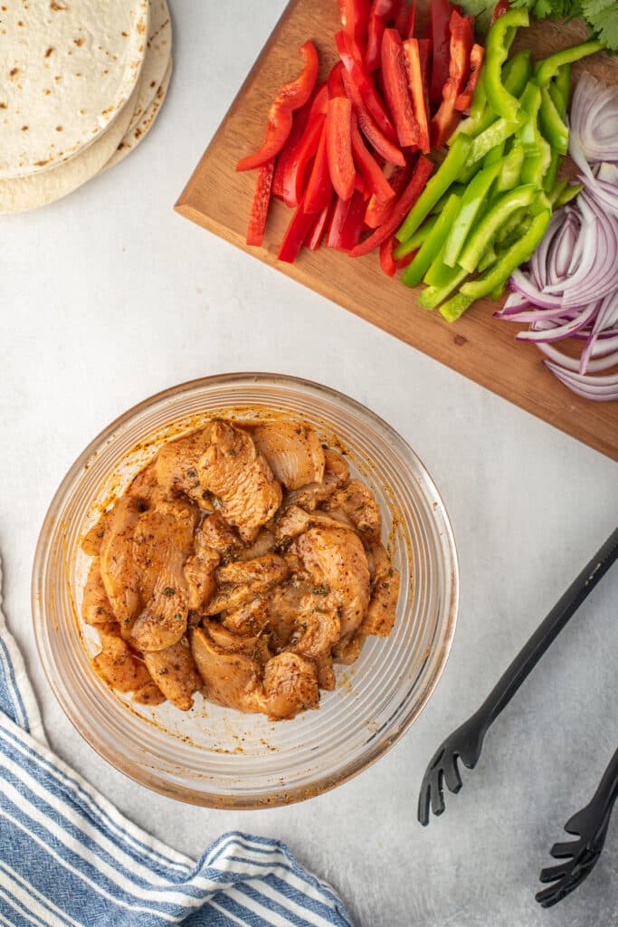 A glass bowl of seasoned raw chicken sits on a counter beside a wooden board with sliced red and green bell peppers, and red onion. Nearby are tortillas, tongs, and a blue-striped kitchen towel.