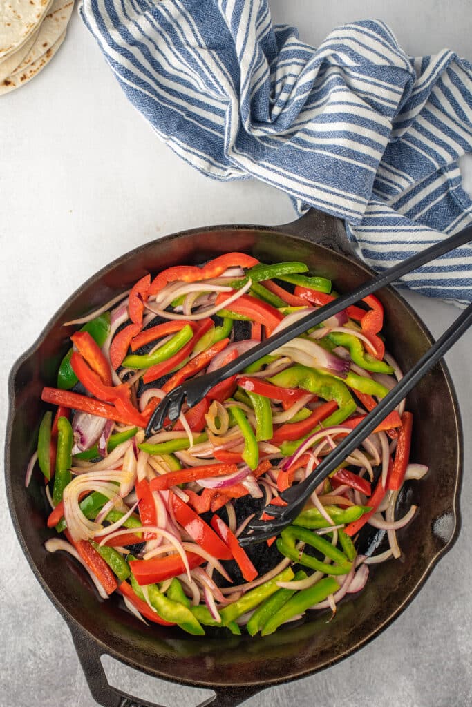 Colorful sliced red and green bell peppers and onions are being sautéed in a cast iron skillet with black tongs. A blue and white striped kitchen towel and some tortillas are nearby on a light surface.