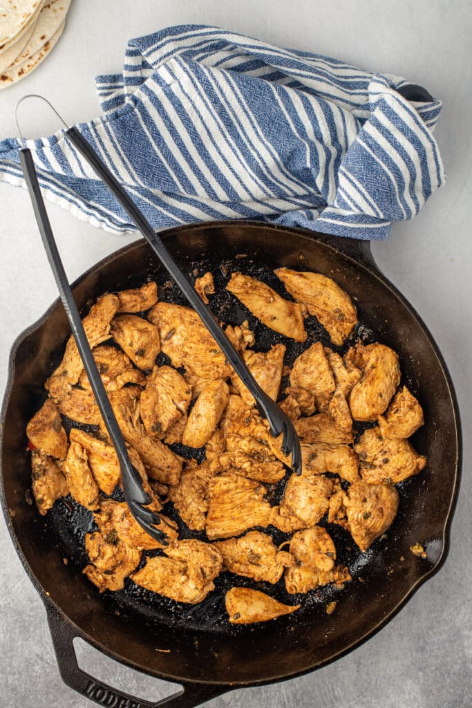 A cast iron skillet filled with cooked, seasoned chicken pieces sits on a gray surface with metal tongs resting inside. A blue and white striped towel and a stack of tortillas are nearby.