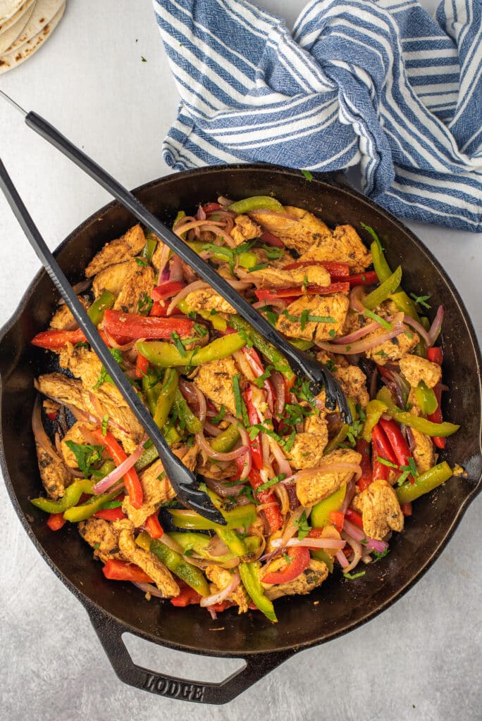 A cast iron skillet filled with cooked chicken strips, red and green bell peppers, and red onions, garnished with herbs. Black tongs rest on the skillet, and a blue-and-white striped towel is nearby.