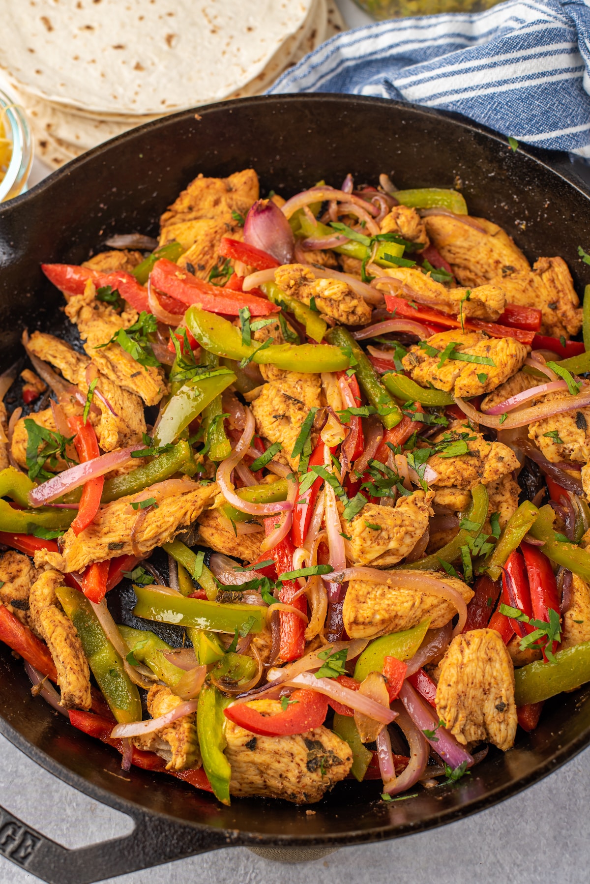 A skillet filled with cooked chicken strips, red and green bell peppers, and onions, garnished with fresh herbs. Flour tortillas are stacked in the background on a blue and white striped cloth.