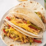 Two soft flour tortillas filled with seasoned chicken strips, sautéed red and green bell peppers, and topped with shredded cheddar cheese on a white plate. A bowl of extra shredded cheese is in the background.