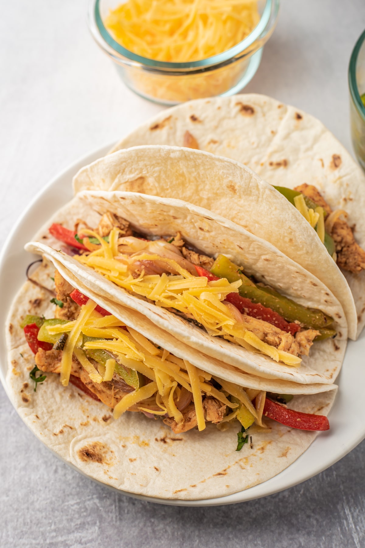 Two soft flour tortillas filled with seasoned chicken strips, saut&eacute;ed red and green bell peppers, and topped with shredded cheddar cheese on a white plate. A bowl of extra shredded cheese is in the background.