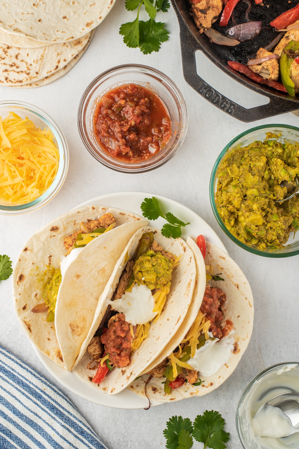 Two soft tacos filled with shredded cheese, sautéed veggies, guacamole, salsa, and sour cream on a plate. Surrounding them are bowls of guacamole, salsa, shredded cheese, tortillas, and a skillet with fajita veggies.