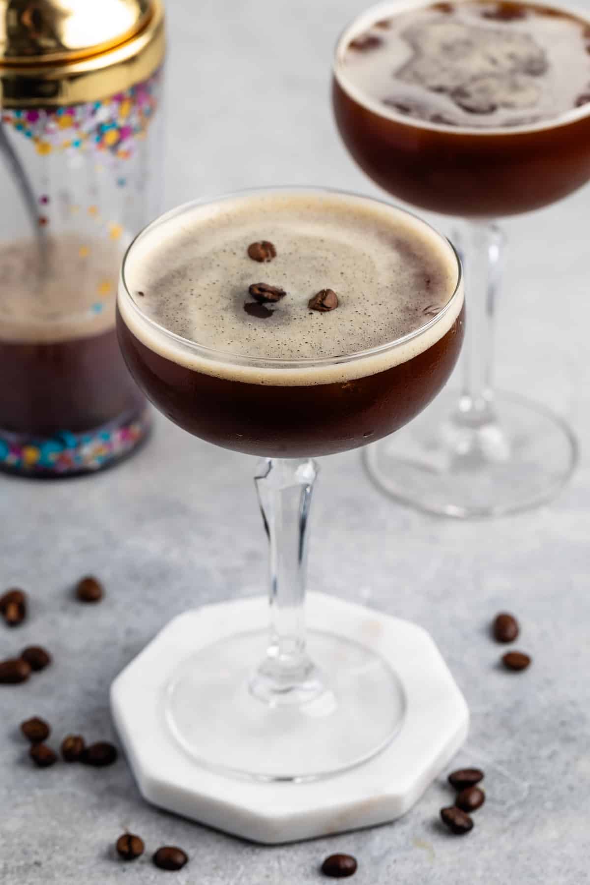 A close-up of an espresso martini in a coupe glass, garnished with three coffee beans, with another glass and a cocktail shaker in the background on a gray surface.