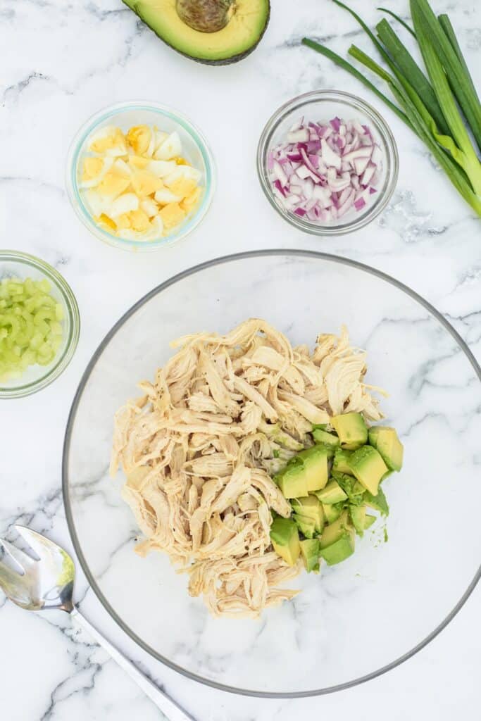 A glass bowl with shredded chicken and diced avocado, surrounded by small bowls of chopped hard-boiled egg, celery, and red onion, with green onions and half an avocado nearby on a marble surface.