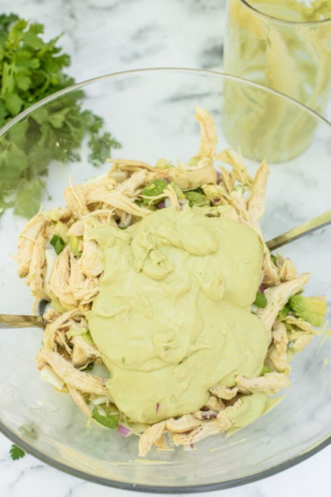 A glass bowl contains shredded chicken, chopped vegetables, and is topped with a creamy green avocado dressing. Fresh cilantro and a glass with dressing are in the background on a marble surface.