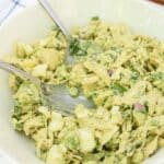 A white bowl filled with creamy chicken salad containing chunks of chicken, hard-boiled eggs, red onion, and green herbs, with two serving forks resting inside. A plate of vegetables is partially visible in the background.
