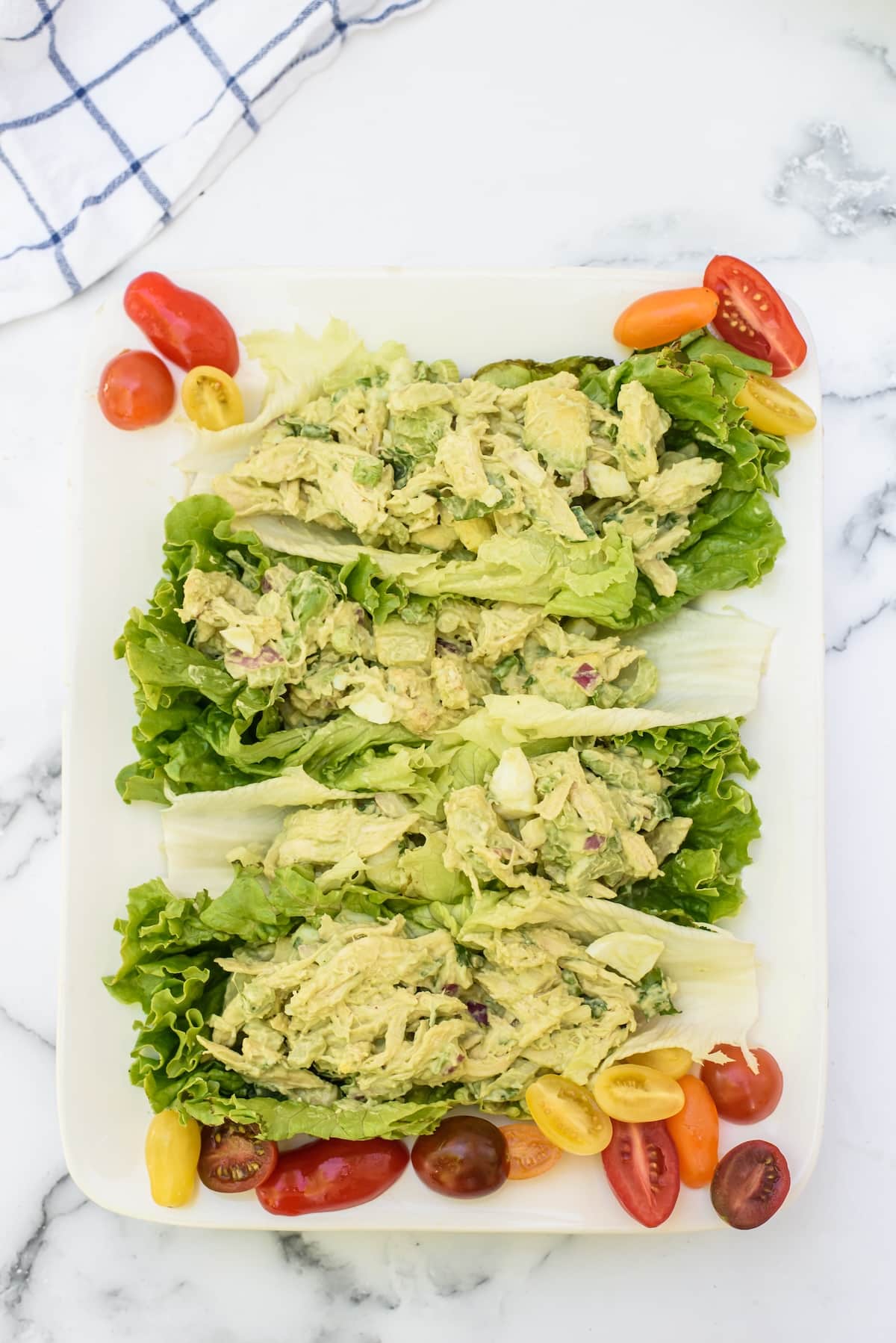 Three lettuce leaves topped with creamy chicken salad are arranged on a white plate, surrounded by colorful halved cherry tomatoes. A blue and white cloth is partially visible in the corner background.