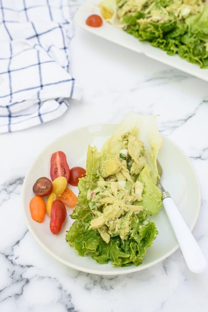 A plate with a lettuce wrap filled with creamy chicken salad, served with colorful cherry tomatoes. A fork rests on the plate, and a striped kitchen towel is in the background.