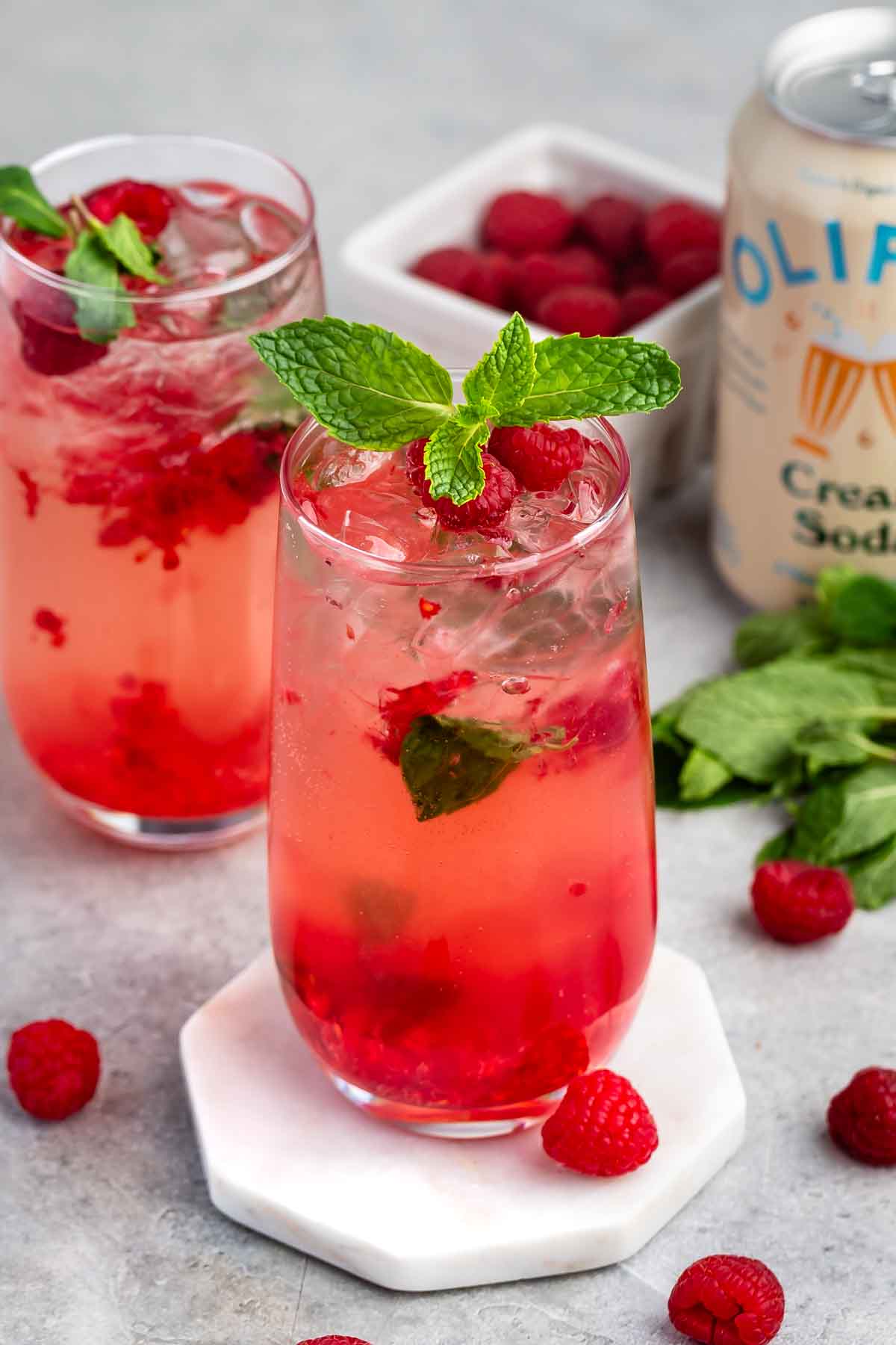 Two tall glasses of a pink raspberry soda garnished with fresh raspberries and mint. Ice cubes are visible in the drinks. Extra raspberries, mint leaves, and a can of cream soda are nearby on a light surface.