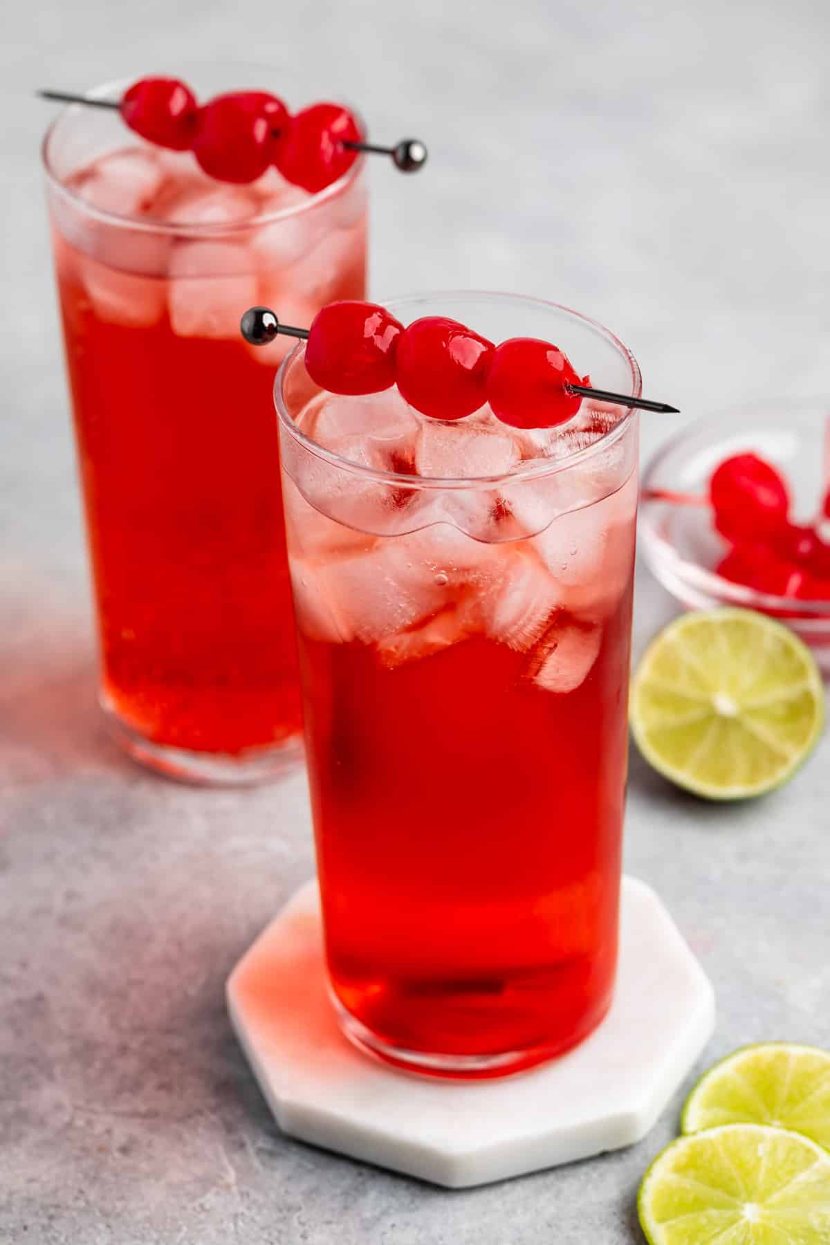 Two tall glasses of bright red iced drinks garnished with skewered maraschino cherries, with a bowl of cherries and sliced limes on a gray surface.
