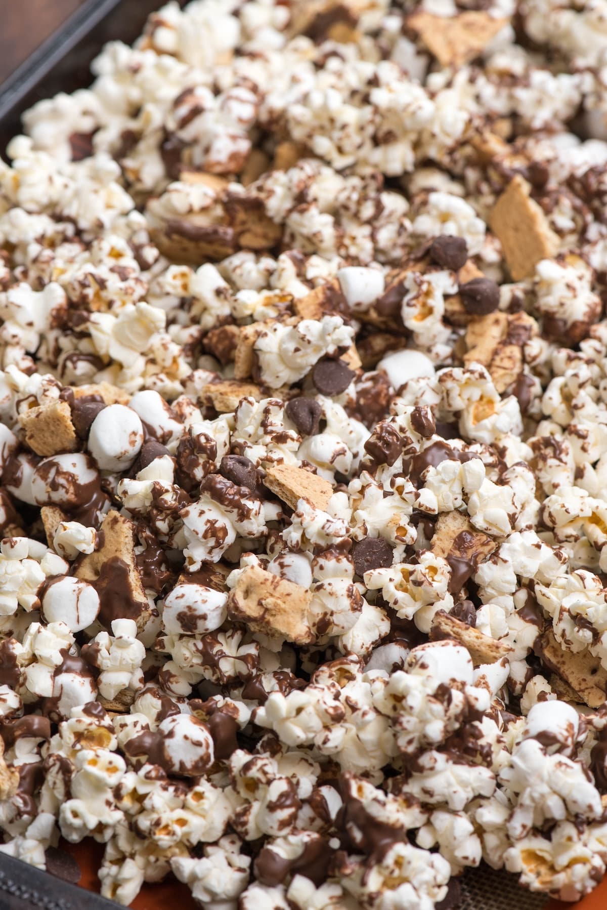 A close-up of popcorn topped with melted chocolate, mini marshmallows, chocolate chips, and broken pieces of graham crackers, resembling a smores snack mix.