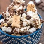A blue bowl with white dots filled with popcorn, mini marshmallows, chocolate chips, and pieces of graham crackers, set on a wooden surface. Some ingredients are scattered around the bowl.