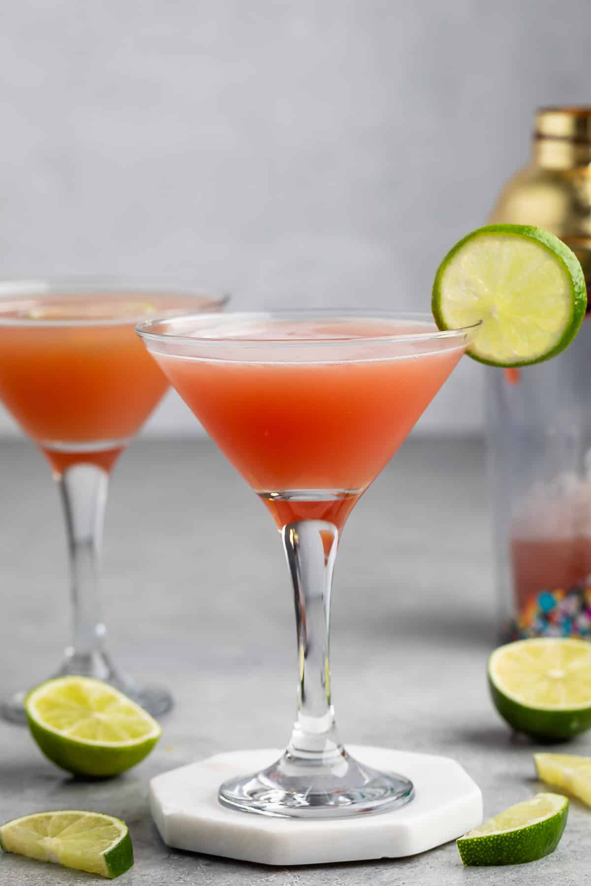 A pink cocktail in a martini glass garnished with a lime wheel, with lime wedges nearby and another identical drink and a cocktail shaker in the background.